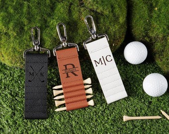 May include: Three personalized golf keychains in black, brown, and white, each with a silver clasp. The black keychain has crossed golf clubs and initials, the brown one has a name, and the white one has initials. Golf balls and tees are also present.