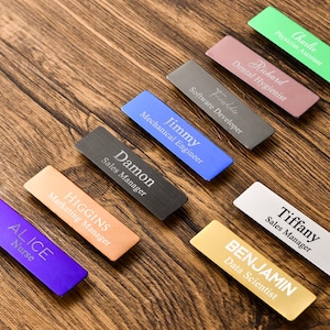 May include: A collection of rectangular metal name badges in various colors including green, pink, gray, blue, black, copper, purple, silver, and gold. Each badge features a name and job title engraved in white or black text on the surface.