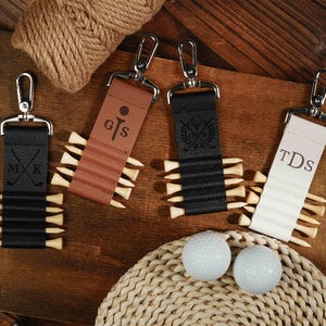 Op de afbeelding: Vier leren golfbalhouders met gepersonaliseerde initialen. Elke houder heeft een metalen clip en een reeks golftees. De initialen zijn "M.K." "G.S." "T.D.S." en een ontwerp van een golfclub en een bal.