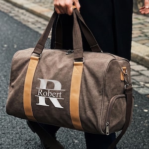 May include: A brown canvas duffel bag featuring two tan vertical leather-like straps and a large white letter R with the name Robert printed horizontally across the center. The bag includes dark brown fabric handles and a side zippered pocket.