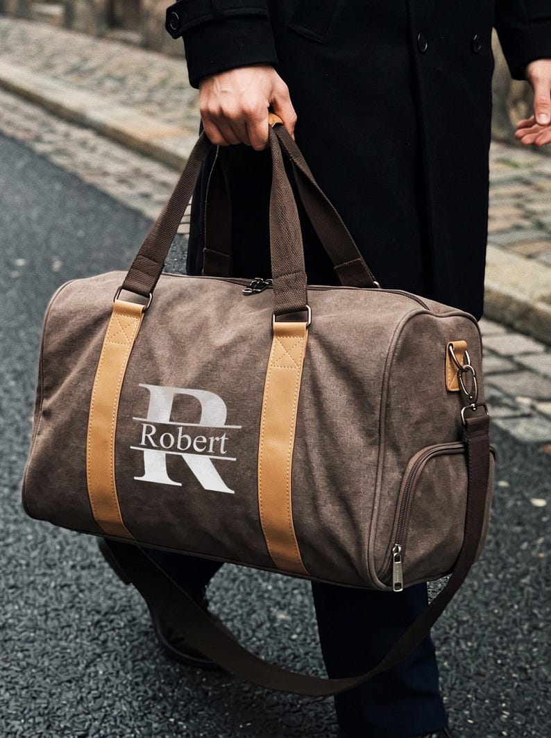 May include: A brown canvas duffel bag featuring two tan vertical leather-like straps and a large white letter R with the name Robert printed horizontally across the center. The bag includes dark brown fabric handles and a side zippered pocket.
