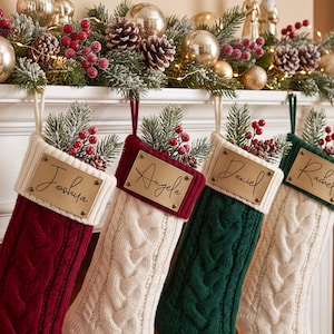 May include: Four cable-knit Christmas stockings in red, cream, and dark green, each featuring a rectangular brass-toned nameplate engraved with the names Joshua, Angela, Daniel, and Rachel, topped with artificial pine branches and red berries.