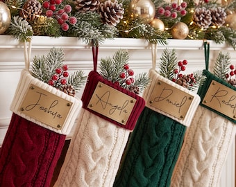 Op de afbeelding: Vier kerstsokken van kabelgebreid materiaal in rood, crème en donkergroen, elk voorzien van een rechthoekig messingkleurig naamplaatje met de namen Joshua, Angela, Daniel en Rachel, versierd met kunstmatige dennentakken en rode bessen.