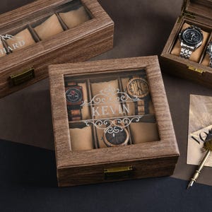 May include: Wooden watch display boxes with glass tops. One box is open, displaying two watches. Another box has the name "RICHARD" etched on the glass. A third box has the name "KEVIN" etched on the glass. The boxes are on a dark surface.