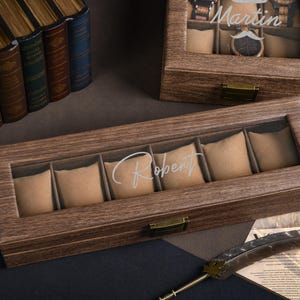 May include: Wooden watch display boxes with glass tops. One box is engraved with the name "Robert", the other with "Martin". The boxes have multiple compartments with beige cushions. A decorative feather and books are in the background.