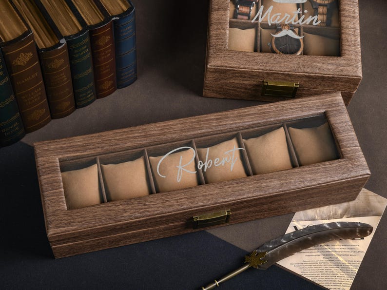 May include: Wooden watch display boxes with glass tops. One box is engraved with the name "Robert", the other with "Martin". The boxes have multiple compartments with beige cushions. A decorative feather and books are in the background.