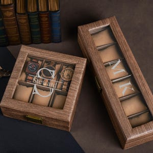 May include: Two wooden watch boxes with glass tops. One box is rectangular and holds six watches, with the name "Steward" engraved. The other box is longer and has the initials "MK" engraved. Both boxes have a brown finish.