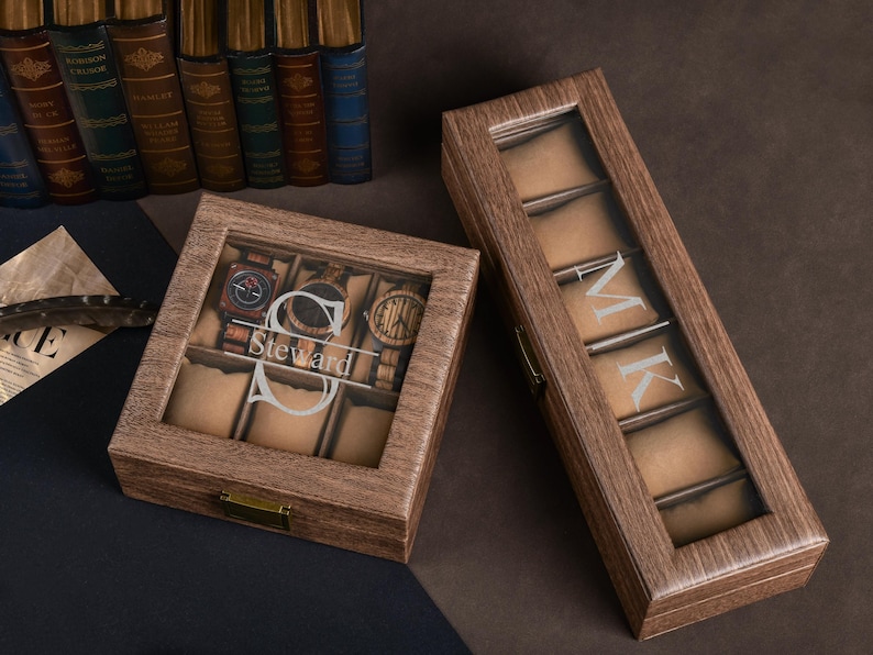 May include: Two wooden watch boxes with glass tops. One box is rectangular and holds six watches, with the name "Steward" engraved. The other box is longer and has the initials "MK" engraved. Both boxes have a brown finish.