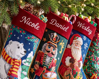 Op de afbeelding: Drie geborduurde kerstsokken met rode fluwelen boorden waarop de namen Nicole, Daniel en Wesley zijn geborduurd. De sokken tonen een witte ijsbeer met een gestreepte sjaal, een notenkraker met een trommel en de Kerstman.
