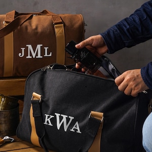 May include: A brown canvas duffel bag with the white monogram JML and a black canvas duffel bag with the white monogram KWA. Both bags feature tan leather-like accents, sturdy handles, and metal hardware, providing a structured design for travel.