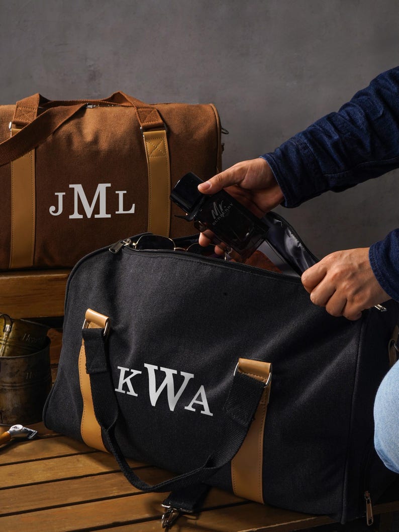 May include: A brown canvas duffel bag with the white monogram JML and a black canvas duffel bag with the white monogram KWA. Both bags feature tan leather-like accents, sturdy handles, and metal hardware, providing a structured design for travel.