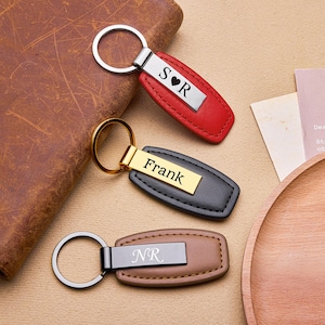 May include: Three personalized leather keychains in red, black, and brown. Each keychain has a metal plate with engraved initials or a name. The keychains are displayed on a light brown surface with a leather wallet and wooden plate.