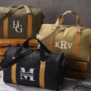 Könnte beinhalten: Drei Reisetaschen aus Segeltuch in Olivgrün, Beige und Schwarz. Jede Tasche hat zwei beige vertikale Riemen, einen Schultergurt und ein weißes Monogramm. Die olivgrüne Tasche zeigt HG, die beige KRv und die schwarze ein großes M mit Michael.