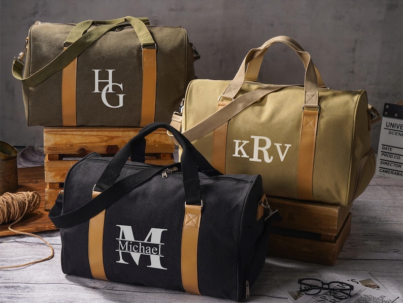 May include: Three canvas duffel bags in olive green, tan, and black. Each bag features two tan vertical straps, a shoulder strap, and a white monogram. The olive bag has HG, the tan bag has KRv, and the black bag has a large M with Michael printed below.