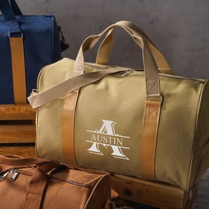 May include: A tan canvas duffel bag with tan webbing handles and tan faux leather accents. The bag features a white graphic print of a large letter A with the name AUSTIN centered horizontally across the letter. A zipper closure is visible.