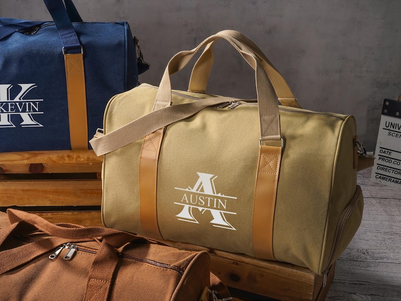May include: A tan canvas duffel bag with tan webbing handles and tan faux leather accents. The bag features a white graphic print of a large letter A with the name AUSTIN centered horizontally across the letter. A zipper closure is visible.