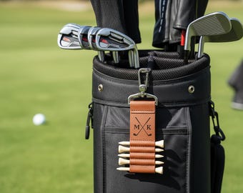 May include: Black golf bag filled with golf clubs. A brown leather tee holder with the initials "MK" and crossed golf clubs hangs from the bag. White golf tees are held in place by the leather holder. A golf ball is visible in the background.
