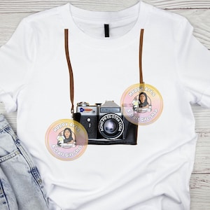 Puede incluir: Camiseta blanca con un diseño de cámara vintage y una correa marrón. La cámara es negra con detalles plateados y una lente prominente. La camiseta también muestra dos logotipos circulares con el texto "Happy Arts & Craft Gift Shop".