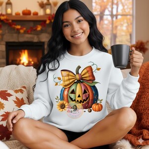 Puede incluir: Sudadera blanca con un diseño de calabaza colorida, un lazo, un girasol y pequeñas calabazas. La calabaza tiene una cara de linterna. Una persona lleva la sudadera y sostiene una taza negra.