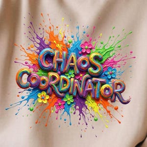 May include: A beige sweatshirt with a colorful graphic that reads "CHAOS COORDINATOR." The design features vibrant, 3D-style lettering and a splash of paint in various colors, including orange, blue, green, and purple, with floral accents.