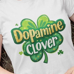 Op de afbeelding: Wit T-shirt met een groene en gouden klaver met de tekst "Dopamine Clover". Het klaverontwerp heeft een gouden glittereffect en steraccenten. Het shirt is gemaakt van een zacht, comfortabel materiaal.