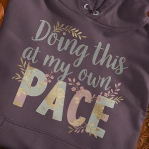 May include: A plum-colored hoodie with the phrase "Doing this at my own PACE" in a decorative font. The word "PACE" is in a pastel color scheme with floral accents, and the hoodie has a drawstring hood.