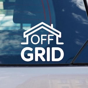 Op de afbeelding: Witte vinyl sticker met de tekst "OFF GRID" onder een huisomtrek. De sticker is op een autoruit geplakt, wat wijst op interesse in een off-grid levensstijl of duurzaamheid.