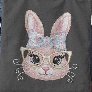May include: A gray tote bag featuring an embroidered bunny face. The bunny has pink fur, large eyes, and wears glasses and a floral bow. The design includes curly white whiskers. The tote bag is made of a durable fabric.