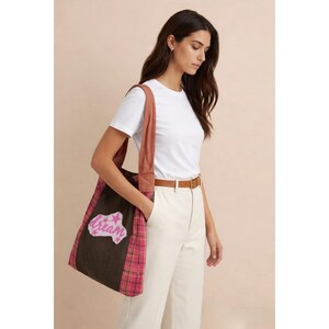 Peut inclure: Un sac fourre-tout marron avec un motif à carreaux roses et une inscription "dream" rose et blanche. Le sac a une sangle marron. La personne porte un t-shirt blanc et un pantalon crème.