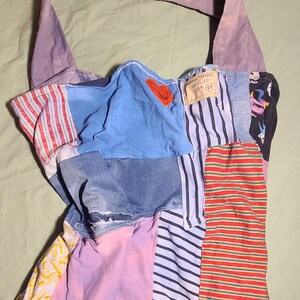 May include: A patchwork tote bag made from various fabrics, including denim, striped patterns, and solid colours. The bag has a light purple handle and a blue section with a red heart applique. The bag is made from recycled materials.