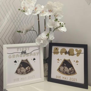 May include: Two white picture frames with black lettering. The frames are decorated with gold lettering that says "First Oh Grandbaby" and "Baby Oh". The frames have a white background and a black mat. The frames are both holding ultrasound pictures and the text "Due May 2025".