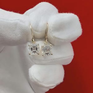 May include: A pair of gold earrings with square-shaped clear gemstones. The earrings are hanging from a white fabric background.