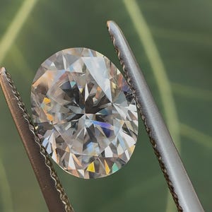 May include: A clear, round, faceted diamond gemstone held in a pair of silver tweezers. The diamond is reflecting light, creating a rainbow effect.