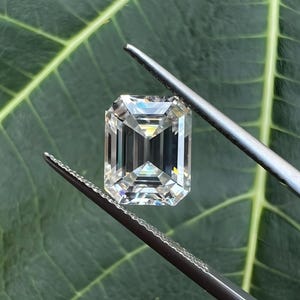May include: A clear, emerald-cut gemstone held by tweezers against a green leaf background.