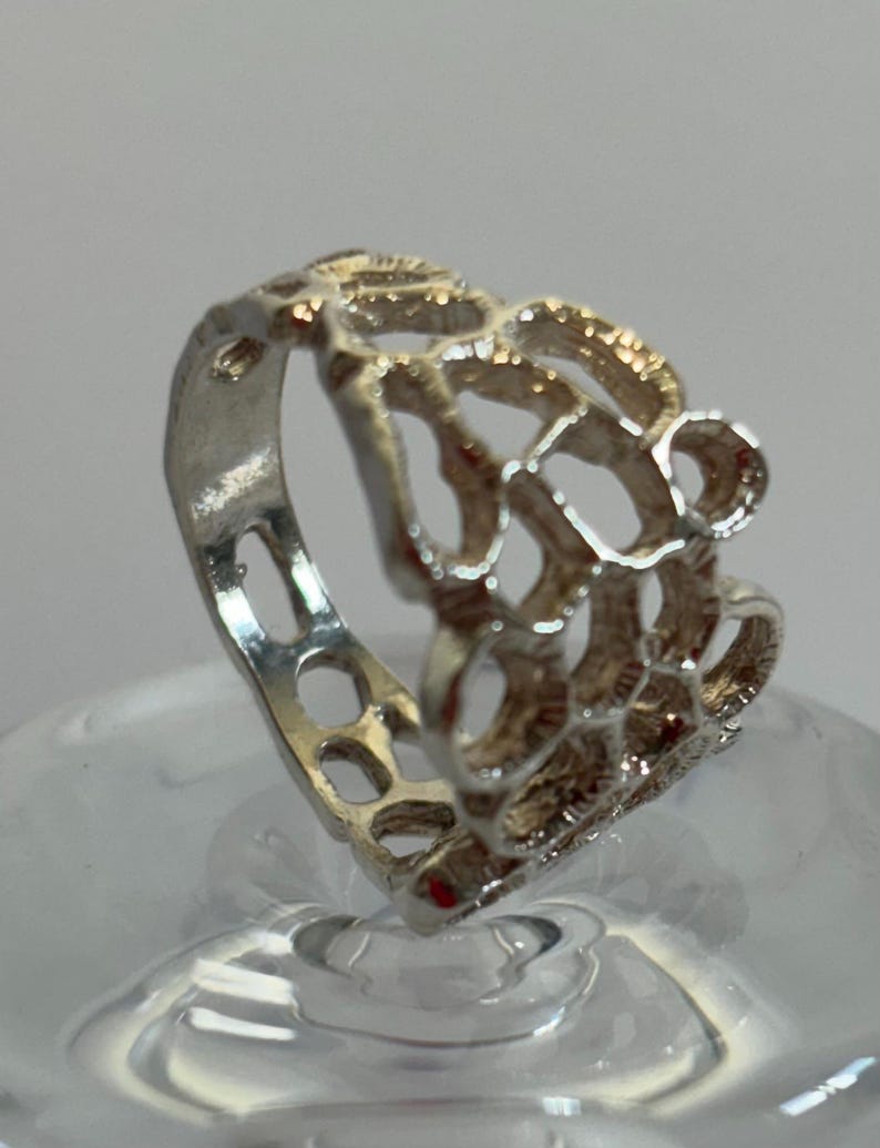 May include: A silver ring featuring a delicate, intricate design. The ring is crafted from a thin band of silver with a pattern of small, interconnected circles and loops. The ring is a unique and stylish piece of jewellery.