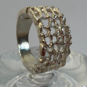 May include: A silver ring with a delicate, intricate design. The ring is made of a thin band of metal with a wide, openwork design that resembles a web or a lattice.