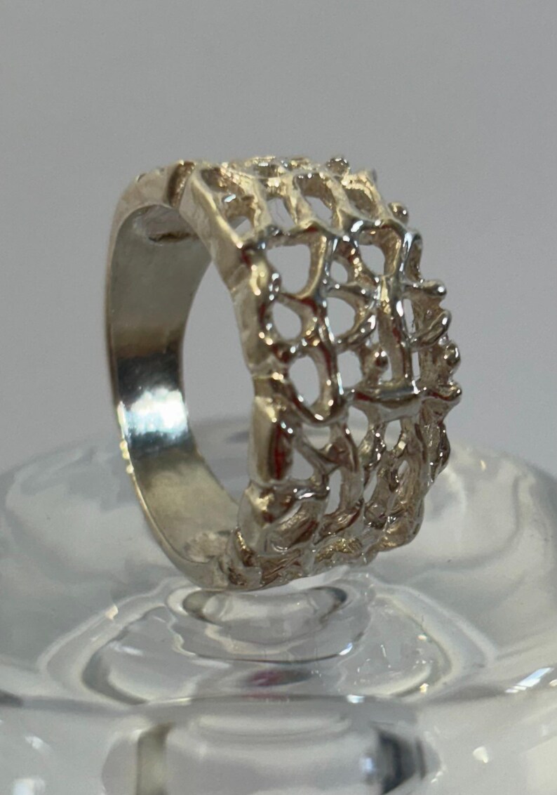 May include: A silver ring with a delicate, intricate design. The ring is made of a thin band of metal with a wide, openwork design that resembles a web or a lattice.