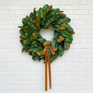 May include: A circular wreath constructed from layered magnolia leaves featuring deep green upper surfaces and brown undersides. The wreath is accented at the bottom center with a long, hanging velvet ribbon in a warm, solid copper-brown tone.