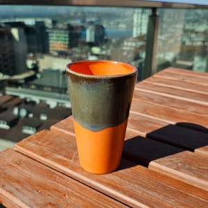 Pode incluir: Um copo de cerâmica feito à mão com uma base laranja vibrante e uma seção superior verde escuro. A borda é um tom de laranja mais escuro. O copo está em uma mesa de madeira com uma vista da cidade ao fundo.