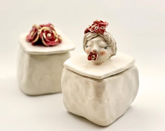 May include: Two off-white ceramic boxes with lids. One lid is topped with a cluster of red and gold roses. The other box features a sculpted face with closed eyes, red lips, and a red flower in its mouth, and a red flower crown.