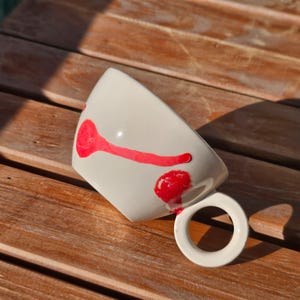 May include: White ceramic mug with a red abstract design. The mug has a round handle and is resting on a wooden surface. The red design appears to be hand-painted.