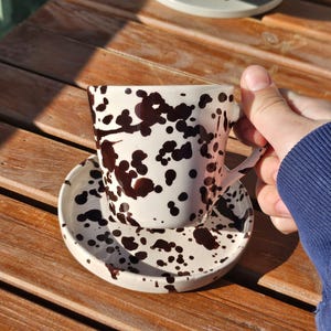 Może przedstawiać: Biały ceramiczny zestaw filiżanki i spodka do kawy z brązowym wzorem rozprysków. Filiżanka ma uchwyt i stoi na pasującym spodku. Zestaw znajduje się na drewnianej powierzchni.