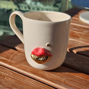 May include: White ceramic mug with a large handle, featuring a red and gold lip design on the front. A small gold accent is placed above the lips. The mug is set on a wooden surface.