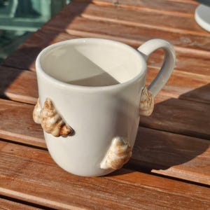 May include: White ceramic mug with a curved handle and seashell embellishments. The mug has three seashells attached to the exterior, adding a coastal touch. The mug is sitting on a wooden surface.