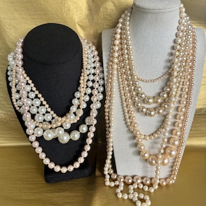 May include: Two pearl necklaces displayed on black and cream-colored busts. The necklaces feature various sizes of faux pearls in white and cream colors. The necklaces are arranged in multiple strands, creating a layered effect. The background is a gold-colored surface.