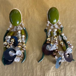 May include: A pair of dangling earrings with olive green oval tops and gold-toned trim. The earrings feature a cascade of clear, gold, and white beads, along with dark green and brown accents. The design includes dark blue and silver circular elements.
