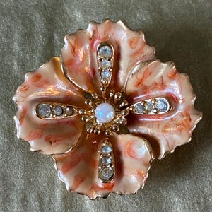 May include: A floral brooch with a four-petal design. The brooch has a peach and gold enamel finish, with clear crystal accents and a central iridescent stone. The brooch is a vintage style.