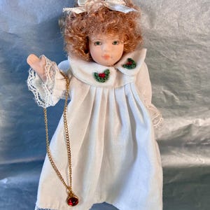 May include: A porcelain doll with auburn hair and green eyes, wearing a white dress with lace trim and a gold necklace with a red pendant. The doll has a white bow in its hair and holly accents on the collar.
