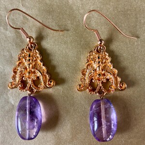May include: A pair of gold-colored earrings with ornate filigree designs. Each earring features a large, oval, purple amethyst gemstone. The earrings have a hook closure.