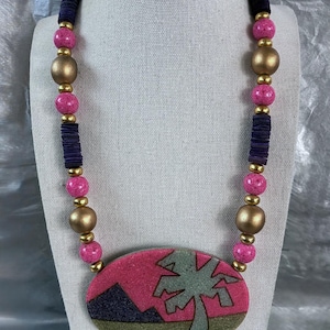 May include: A statement necklace featuring a pink and gold pendant with a palm tree design. The necklace has pink, gold, and navy blue beads. The pendant is an oval shape with a pink background and a green palm tree.